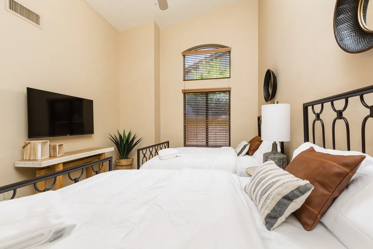 A cozy bedroom featuring two beds with decorative pillows, a television, an indoor plant, and a window with blinds. The space is designed for relaxation, entertainment, and flexible sleeping arrangements.