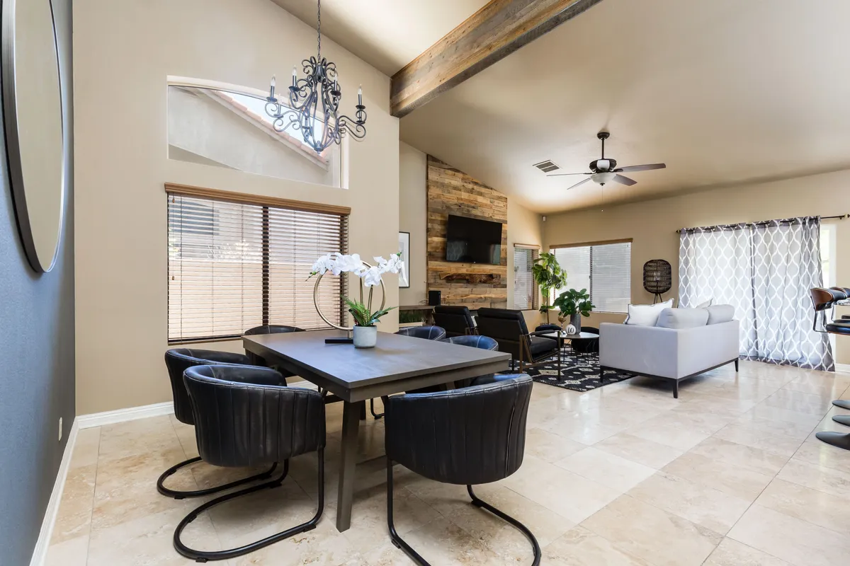 A modern, inviting dining area with a large table, black chairs, a stylish chandelier, a wooden accent wall, and expansive windows leading into a comfortable seating area. The space is designed for social gatherings and collaborative meals.