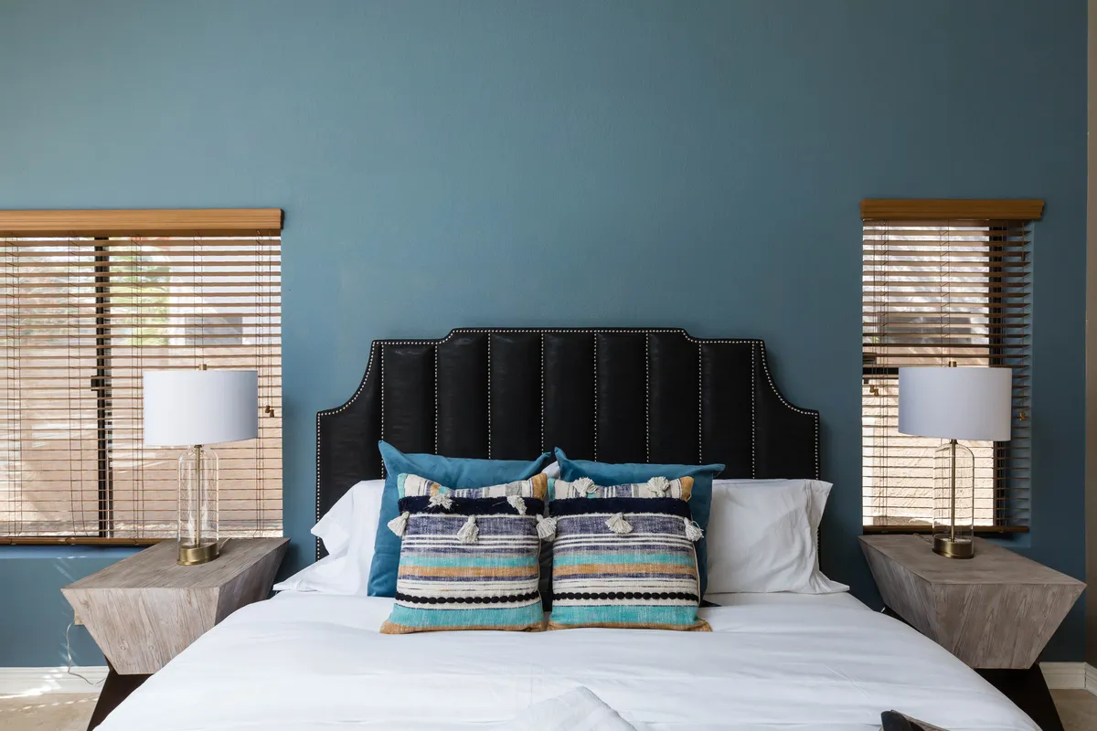 A tranquil bedroom with a bed adorned with decorative pillows, nightstands with lamps on either side, and windows with blinds letting in soft natural light. The setting is peaceful and perfect for unwinding after a busy day.