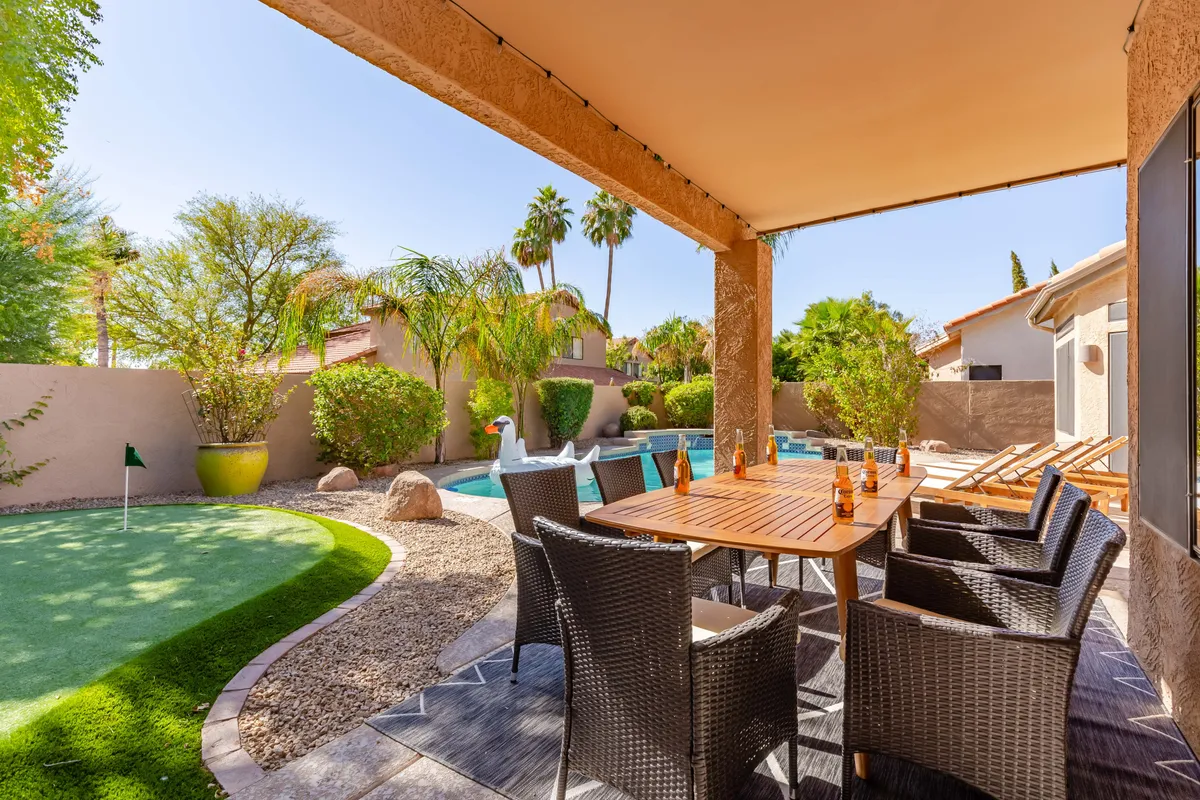 The image showcases the backyard oasis of a Scottsdale single-family home, featuring a sparkling saltwater pool surrounded by lush greenery. A spacious outdoor dining table invites guests to enjoy meals alfresco, while a manicured putting green offers space for golf practice. The setting is infused with a cheerful, leisurely vibe, perfect for sunset dinners, poolside lounging, and friendly putting competitions.