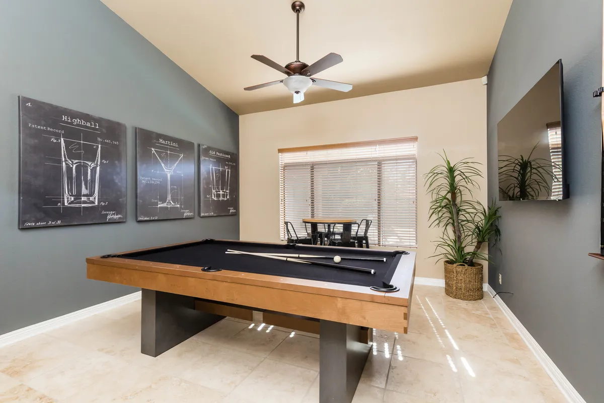 A lively entertainment lounge featuring a classic pool table, cocktail-themed artwork, indoor plant, window with blinds, and a nearby dining table. The setup is casual and fun, offering space for games, drinks, and relaxed socializing.
