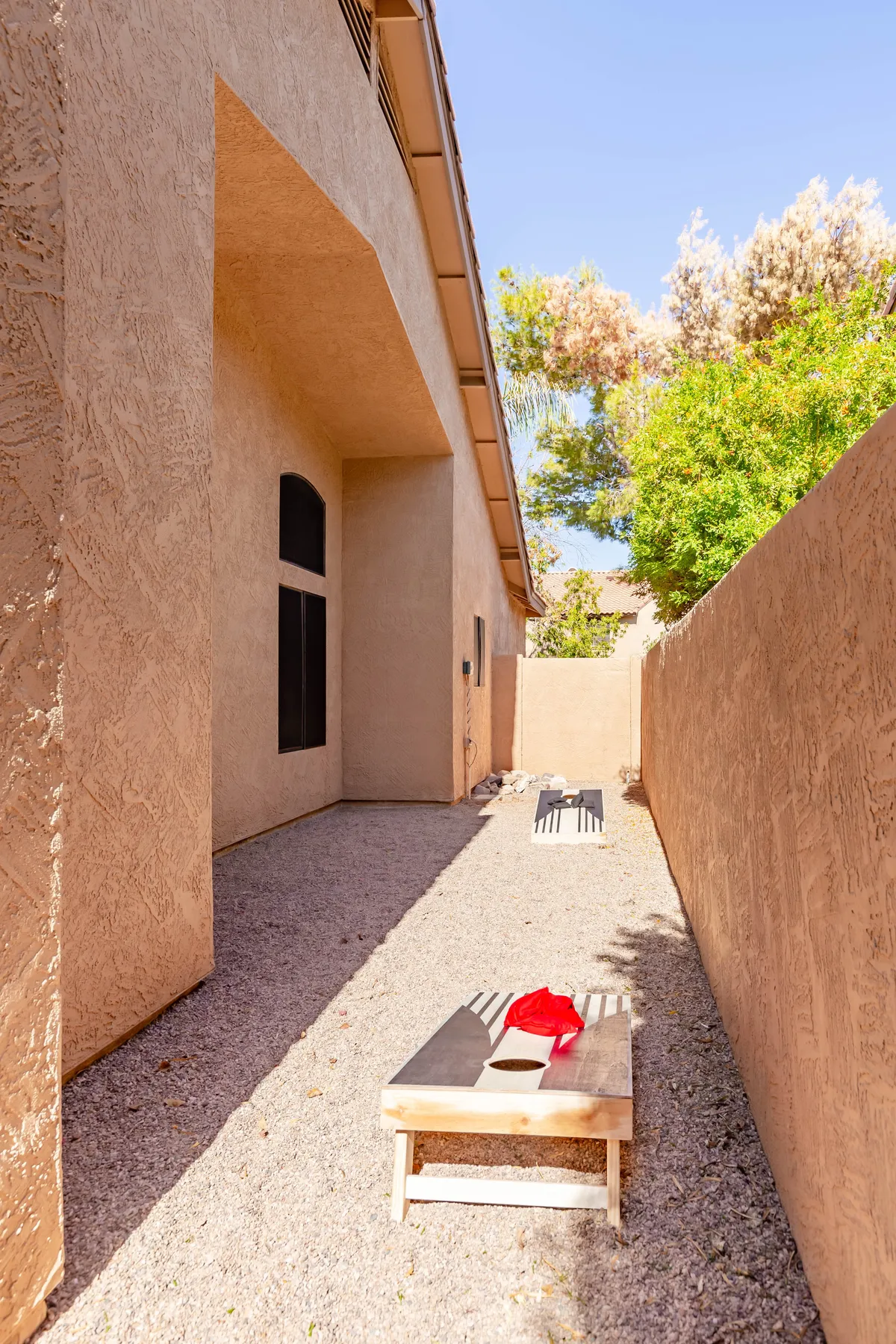 A dedicated games area features two bean bag toss boards set on a gravel pathway, with sunlight casting inviting shadows. Friends are gathered, playing and socializing, creating an atmosphere of friendly competition and laughter. The area is framed by greenery, further tying it to the lush outdoor setting described in the section.